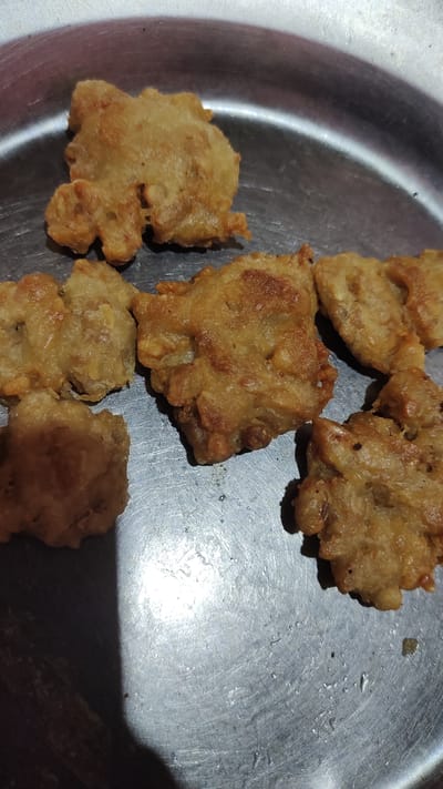 Six pieces of fried food are arranged on a circular metal plate. The food appears to be savory, with a golden-brown, crispy exterior. The plate has a brushed metal finish with visible circular scratches, and a strong light source casts a bright reflection on the left side of the plate, creating a sharp shadow to the left of the food items. The background is a blurry, metallic surface consistent with a dining table. The scene is likely indoors, given the lighting and indoor setting. There are no people or animals visible in the image. The exact location in Palangka Raya, Indonesia is not discernible from the provided image alone.