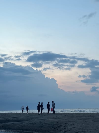 The scene depicts a beach environment in Angkleng Barat, Indonesia. Several people are present, silhouetted against the sky and the sea. The beach appears to be dark sand. The sky is partly cloudy, with a mix of light blue and darker, possibly stormier clouds. A streak of pinkish-orange suggests either sunrise or sunset. The ocean meets the beach with visible waves. People are walking on the beach.