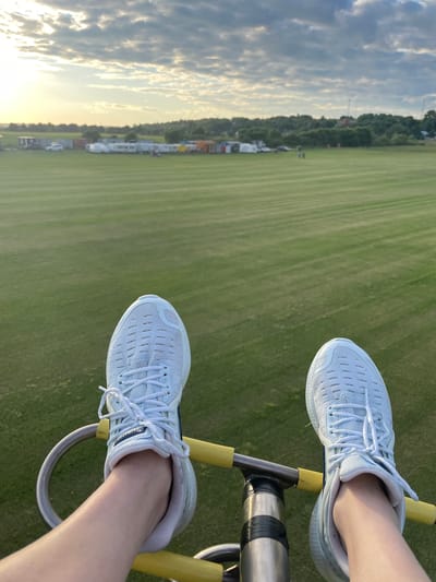 The image shows a first-person perspective of someone's feet wearing white athletic shoes, positioned on a metal and yellow-bar apparatus. The feet are against a backdrop of a vast, well-maintained green field with striped patterns from mowing. In the distance, a row of mobile homes or trailers is visible, along with some trees and structures. The sky above is partly cloudy with sunlight breaking through, indicating it might be late afternoon or early morning. The location is identified as Турово, Russia.