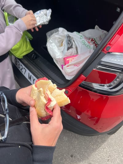 The image shows two individuals outside a red car, likely in La Aldea de San Nicolás, Spain. One person is unwrapping food in aluminum foil, while the other holds a half-eaten sandwich with cheese and salami. The car's trunk is open, revealing grocery bags inside. The number plate of the car visible is '3327 MY'. The environment appears to be an outdoor parking area or roadside. One of the people is wearing a black hoodie, while the other is wearing a light purple long sleeve.