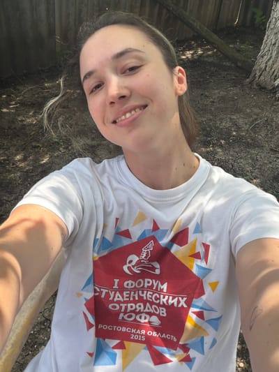 A young woman with light brown hair pulled back smiles for a selfie. She is wearing a white t-shirt with a red, yellow, and blue graphic design and text. The text on the shirt reads "I ФОРУМ СТУДЕНЧЕСКИХ ОТРЯДОВ ЮФО РОСТОВСКАЯ ОБЛАСТЬ 2015". She is seated outdoors in what appears to be a yard, with dry, cracked ground and a wooden fence visible in the background. A tree trunk is also present to the right. The lighting suggests it is daytime.