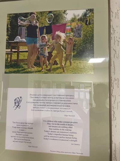 The image shows a framed print. The print contains a photograph of a woman and three children outside. The woman is blowing bubbles while lying on a wooden platform or bench. The children are interacting with the bubbles. Laundry is hanging on a line in the background, with trees visible beyond the laundry.
The right side of the print includes text in three languages (likely Russian, English, and another). The text is attributed to Sri Chinmoy. There is also a small butterfly-like symbol below the Russian text. The text is about children making constant progress and how dreams become realities. At the very bottom right corner, it mentions "Рисунок Птиц-Душ выполнен Шри Чинмоем".
The location of Sanatoriya Yumatovo Imeni 15-Letiya Bassr, Russia is not discernable from the image alone.