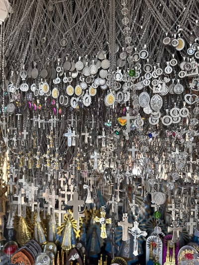 The image displays a dense collection of silver necklaces with varying pendants. A multitude of round, oval, and cross-shaped pendants are visible, interspersed with beads and other embellishments. The arrangement is closely packed, creating a textured effect. Religious iconography is prominent, with numerous crucifixes. The necklaces appear to be arranged in a retail setting, possibly for sale.