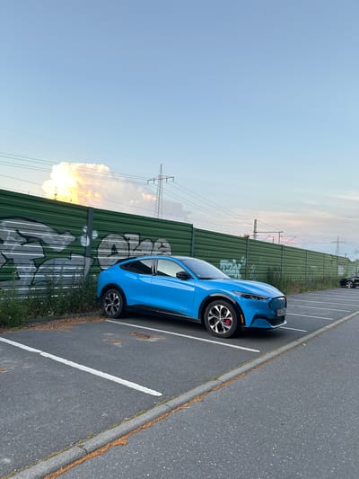 A bright blue Ford Mustang Mach-E is parked in an empty parking lot, facing right. The car is in the foreground and occupies the left half of the frame. Behind the car is a tall, green sound barrier covered in graffiti. In the distance, power lines and a pylon are visible against a pale blue sky with some clouds. The parking lot is paved with asphalt and has white parking lines. The overall scene is an outdoor urban environment.