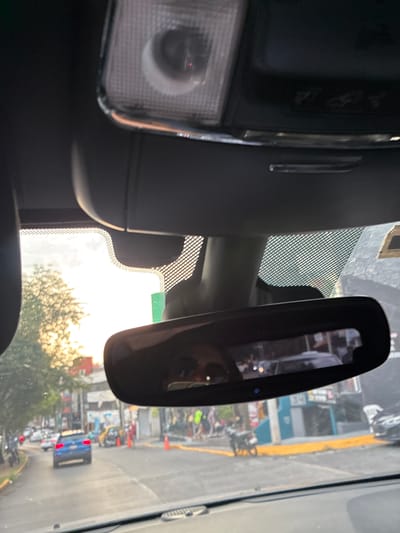 A view from inside a vehicle shows a street scene in Atizapán de Zaragoza, Mexico, as reflected in the rearview mirror. The reflection shows a street with buildings, pedestrians, vehicles, and trees. The street appears busy, with possible construction or road work indicated by traffic cones. The sky is visible through the windshield, suggesting it's daytime. A portion of the vehicle's interior, including the dome light and rearview mirror, is also visible.
