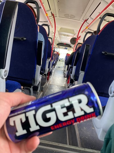 A hand holds a can of "Tiger Energy Drink" in the foreground of an image taken inside a bus. The bus interior features rows of dark blue upholstered seats with black headrests and silver frames. Red and white grab bars are visible along the ceiling. The floor of the bus is dark gray. Several passengers are seated, facing forward. One passenger in the foreground appears to be wearing a light blue jacket. The background shows the aisle of the bus, with more rows of seats and passengers. The ceiling has "Mercedes-Benz" branding. The location is identified as Bielany Wrocławskie, Poland, implying this is a public transportation vehicle.