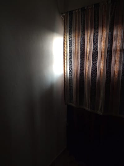 A dimly lit interior scene shows a textured wall on the left, illuminated by a bright, harsh light source casting a strong, elongated glow. The light creates a sharp gradient across the wall, with a significant portion of the left side in deep shadow. To the right, a set of striped curtains hangs, featuring vertical bands of dark blue, light tan, and orange, with an intricate pattern within some of the darker stripes. The curtains obscure the view beyond them. The bottom of the image is dominated by dark, indistinct shapes, suggesting furniture or floor. The overall atmosphere is one of shadow and controlled illumination. This scene is located within the Limuru Central ward, Kenya.