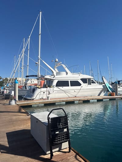 The scene is a marina in La Paz, Mexico, under a clear blue sky. Several sailboats and a larger white motor yacht are docked along a wooden pier. The water is calm with visible reflections. A black metal gangway or ladder is on the pier next to a gray concrete box. Details of masts, rigging, and electronic equipment are visible on the boats.
