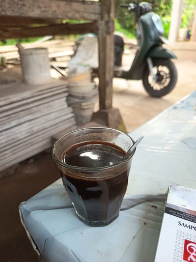 A glass of dark coffee sits on a light-colored, plastic-covered table in what appears to be an outdoor, covered area. A spoon is in the coffee. To the right of the coffee is a partially visible cigarette pack. In the background, a dark green scooter is parked. Behind the scooter, wooden planks are stacked, and buckets and bags are visible. The environment suggests a casual, possibly utilitarian setting, such as a workshop or a rural roadside stop. The lighting indicates it is daytime. The location is identified as Rayeukmatang, Indonesia.