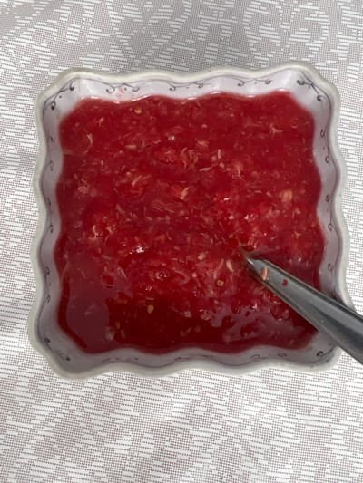 An overhead, close-up view displays a white, square-shaped ceramic bowl with scalloped edges and a subtle dark blue or grey decorative pattern along its rim. The bowl contains a bright red, viscous substance with visible lighter-colored fibrous fragments and small seeds, consistent with a chunky sauce, relish, or jam. A metallic, silver-toned spoon is partially submerged in the substance, its handle extending diagonally towards the bottom right of the frame. The bowl rests upon a white or light-colored tablecloth featuring a repeating, subtle geometric pattern. No individuals are visible. The scene is located in the city of Пугачёво, Russia.