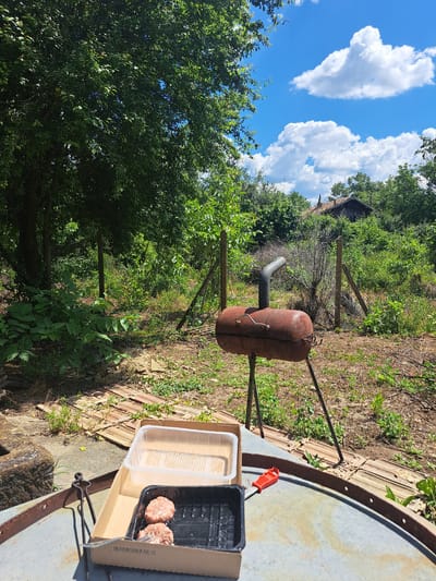 In the city of Kardam, Bulgaria, a rustic outdoor scene unfolds under a partly cloudy blue sky. In the foreground, a metal structure, possibly a repurposed barrel or basin, serves as a makeshift table. On it rests a cardboard box containing a black plastic tray with two raw hamburger patties, alongside a clear plastic container. To the right, a small red utensil with prongs lies on the edge of the metal surface.
Behind this setup, a rusty, old-fashioned barrel-shaped barbecue grill, perched on three legs, stands ready. A bent metal pipe protrudes from its top as a chimney. The grill is positioned in a slightly overgrown yard, with a wire fence and wooden posts marking a boundary in the middle ground. Beyond the fence, dense green foliage and trees dominate the landscape, with the roofline of a dilapidated building barely visible through the greenery. The ground is a mix of dirt, grass, and scattered debris, including what appears to be broken roof tiles or bricks. The overall impression is one of a neglected but functional outdoor cooking area in a rural setting.