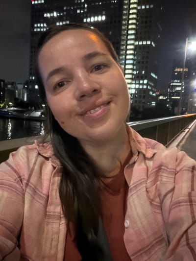 A young woman with dark hair and a mole on her left cheek is taking a selfie at night on a pedestrian bridge in Koto, Japan. She is wearing a pink plaid shirt over an orange shirt. The background shows a body of water with reflections of lights, and tall buildings with illuminated windows. A street light is visible on the right side of the image.