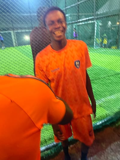 In Malete, Nigeria, two individuals are photographed on an indoor football pitch. The foreground is dominated by the back of a person wearing an orange jersey and shorts. To the right and slightly behind, another person in an orange jersey and shorts is visible. This person is smiling broadly, revealing well-defined teeth. The background shows a green artificial turf, a white net enclosure, and other figures in the distance, some appearing to be playing football. The lighting suggests it is evening or night.
