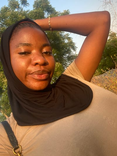 A young woman wearing a black headscarf and tan shirt lies on her back outdoors, with trees visible in the background. Her arm is raised, adorned with a gold bracelet. The light reflects off her face and skin.