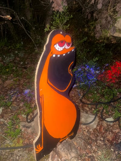 The image shows a cartoonish, Halloween-themed wooden lawn decoration of an orange and black cat with bulging eyes and a toothy grin, situated among ground cover and foliage. Red and blue string lights are visible in the background. The overall environment suggests an outdoor setting at night or in low light. The location in the city of Étréchy, France, is not identifiable in the image.