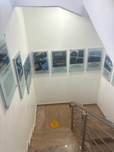 This image captures an indoor setting, likely a stairway or ramp area in a museum, educational facility, or public building, located in Gwaska, Nigeria. The scene is brightly lit, showcasing white walls and a smooth, light-colored floor, contrasting with a section of mottled brown and grey marble-patterned wall next to a modern stainless steel railing on the right.
The primary subjects are multiple glass plaques affixed to the white wall on the left, displaying images and text, mostly related to aviation. Several similar glass plaques also lie flat on the floor, continuing the display downwards. No people are visible in the scene.
Notable details include the geometric lines of the architecture and the clean, contemporary aesthetic of the display. The wall plaques are arranged vertically, with images of aircraft and aircraft cockpits. The plaques on the floor appear to have more textual information. On the marble-patterned wall next to the stairs, a yellow circular sign with black text is affixed.
Visible text from the plaques and signs includes:
* "LUFTHANSA AIRLINES"
* "AER LINGUS"
* "THE COCKPITS OF"
* "RAPTORADAS FIGHTING FALCON" (likely referring to F-22 Raptor and F-16 Fighting Falcon)
* "THE JETSTAR AIRWAYS"
* "500" (possibly related to an aircraft model, e.g., B-52 Stratofortress, which is typically accompanied by images of aircraft engines/turbines as seen in one plaque)
* "THE NORTH AMERICAN ROONE" (possibly referring to the North American B-25 Mitchell bomber, or a similar historical aircraft)
* A yellow sign reads "MIND YOUR STEP".
