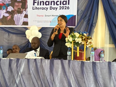 This image depicts an indoor financial literacy event, likely a conference or seminar, held at Unguwan Dosa, Nigeria. The central focus is a woman, presumably a speaker, standing behind a draped table and addressing an unseen audience. She is dressed in a black blazer over a red top, with a dark head covering, holding a microphone in her right hand and gesturing expressively with her left. Her hands are adorned with rings and bracelets.

Seated to her left at the table is a man in a dark suit and tie, looking down at documents and a laptop. Another man's head is partially visible behind him. On the far right, a woman in a purple hijab is partially visible. The table is covered with a pleated, translucent purple-blue fabric. Objects on the table include a laptop, various papers, two bottles of "Swan" brand water, and a floral arrangement featuring white and blue artificial flowers. A cluster of white balloons can be seen to the left of the speaker.

In the background, a large banner announces "Financial Literacy Day 2026" with the theme "Smart Money Talks." The banner also displays contact information: "TOLL FREE HELP DESK: 080063424357," "HELP DESK E-MAIL: helpdesk@ndic.gov.ng," and social media handles such as "@NDICNigeria." An image of children in a classroom, with the text "Smart Money Talks," is also part of the banner's design. The venue's background is decorated with blue and light-colored sheer drapes, suggesting a formal or semi-formal setting. The event appears to be organized by or in collaboration with "NDIC."