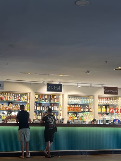 An interior view depicts a modern bar area with two male patrons standing at the counter and a partially visible bartender. The bar's front is a prominent teal-green with vertical ridges, subtly illuminated. The bar counter surface is dark brown or black, equipped with beer taps on the left and various bar tools. Behind the counter, four vertically stacked, illuminated shelving units display a wide array of liquor, wine, and spirit bottles.
Two dark rectangular signs hang from the ceiling above the back bar, labeling sections as "Cocktail" (center-left) and "Beverages" (right); a partial sign reading "ks" is visible on the far left. The ceiling is light gray, featuring multiple recessed circular lights and track lighting with spotlights. Small, dark, ceiling-mounted sensors or cameras are also visible. The floor is dark and appears to be polished.
Two men are standing with their backs to the viewer at the bar counter. The man on the left wears a black short-sleeved t-shirt, light-colored shorts, and dark flip-flops. The man on the right wears a black short-sleeved t-shirt with white graphics on the back (including "HUGO"), black shorts, and black flip-flops. He carries a black shoulder bag and is captured in the act of drinking from a glass. A bartender, mostly obscured, is seen bent down behind the central section of the bar.
No visual information within the image confirms the specified location as Selçuk, Turkey.