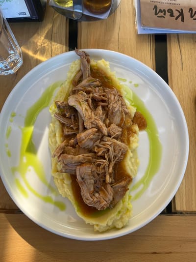 A top-down shot reveals a plate of food on a wooden surface. The plate is round and white, containing a bed of mashed potatoes topped with shredded meat and a sauce. A green drizzle garnishes the plate. Parts of a paper menu, glass, and a black object are visible on the table. The image was taken in Slokoshtitsa, Bulgaria.