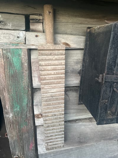 The image shows a close-up, top-down view of rustic, weathered wooden surfaces, likely in an outdoor setting such as a shed or an old barn in Пярэжырскі сельскі Савет, Belarus. Dominating the foreground is a wooden washboard with distinct parallel ridges, its handle made of a lighter, round piece of wood attached with nails. Below the washboard, a wider, greenish-painted wooden plank is visible, showing signs of age and wear. In the upper portion of the image, a dark, rectangular object, possibly a metal chest or box with a lid and latch, rests on wooden planks. The scene is dimly lit, suggesting it might be indoors or during a less sunny part of the day. The overall impression is one of historical preservation or an artifact display, highlighting old tools and construction methods. No people are visible, and the focus is entirely on the objects and their aged textures. The location cues, such as the style of construction and the washboard, suggest a rural Belarusian context.