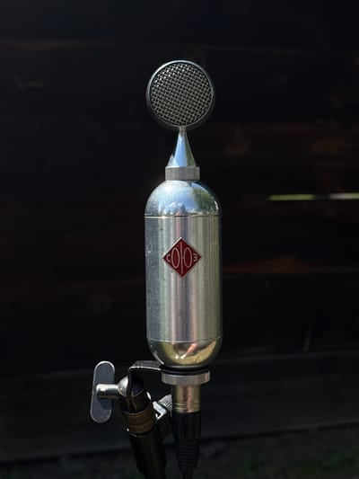 The image shows a close-up, vertical, eye-level shot of a vintage silver microphone mounted on a black microphone stand. The microphone has a rounded grille at the top, followed by a conical section and a cylindrical body. A diamond-shaped emblem with red detailing is visible on the cylindrical body. The microphone stand has a black articulated arm with a metal T-shaped adjustment knob. The background is a dark, textured wooden surface, likely part of a building, with some hints of green foliage at the bottom suggesting an outdoor setting. The lighting is natural and bright, casting subtle shadows on the microphone. There is no indication of the specific location within the city of Shushenskoye, Russia, or any people present in the image.