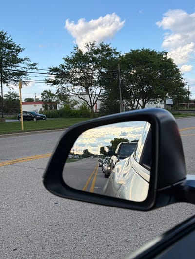 The image shows a view from a car looking at the side mirror. The side mirror reflects a road with several cars in traffic, under a cloudy sky. In the foreground, the car's gray asphalt road is visible. In the background, there are trees, a building, and a street. The scene is set in Mundelein, United States of America.