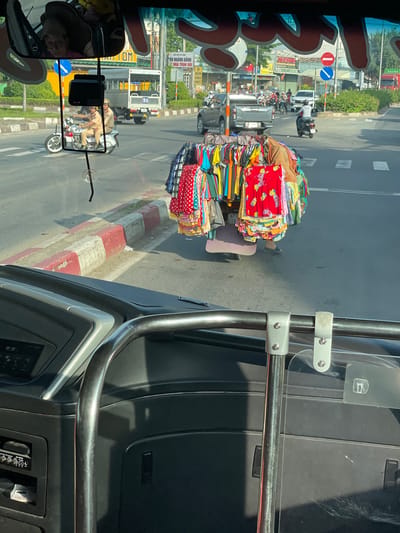 This image, captured from inside a vehicle, shows a busy street scene in Trảng Bàng, Vietnam. In the foreground, the dashboard and interior of the vehicle are visible, including a metal bar with a clear plastic shield attached. Outside the vehicle, a vendor is seen pushing a cart laden with colorful clothing. The cart is covered in a variety of garments, including patterned dresses and shirts. Behind the vendor, two police officers on a motorcycle are visible, as well as a white truck and other vehicles on the road. The street is lined with buildings and vegetation, suggesting an urban or semi-urban environment. The overall scene depicts a typical day in Trảng Bàng, with commerce, law enforcement, and daily life unfolding simultaneously.