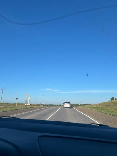 The image is taken from the perspective of a vehicle driving on a two-lane asphalt road in Ishmurzino, Russia. The road is bordered by grassy shoulders and a gentle slope on the right. In the distance, a white car is driving ahead in the same direction. Several power poles with overhead wires are visible along the side of the road. A tall radio tower stands in the middle ground on the right. The sky is clear blue, with a few wispy clouds. The foreground shows the interior of the vehicle, specifically the dashboard.