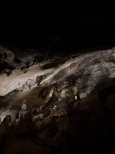 The image shows a dimly lit interior view of the Caves of Drach in Manacor, Spain. The foreground is dominated by the rough texture of cave walls, displaying a mixture of light and dark gray tones. Stalactites and stalagmites are visible on the walls. At the bottom right, a few individuals appear to be part of a tour group, faintly illuminated and partially obscured by the cave formations. The cave's irregular surface is the primary focus, showcasing the natural formations within the caverns.
