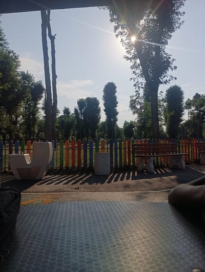 The image depicts an outdoor scene in a park, likely during the daytime, given the bright sunlight and clear sky. The sun is shining intensely, casting long shadows from the trees and a colorful, segmented fence that runs across the middle ground. The fence is made up of vertical posts painted in alternating colors of blue, green, orange, and red.
In the foreground, a textured surface, possibly a table or the ground, fills the bottom portion of the frame. To the left, a part of a person's leg is visible, suggesting someone is sitting or resting.
Behind the fence, park furniture is visible, including a uniquely shaped light-colored seating structure with a metallic inset on the left, and a wooden bench with stone supports on the right. A cylindrical trash receptacle is also present near the bench.
The background is dominated by lush green trees of various sizes, with tall, slender trees on the left and a larger, more dense tree with visible leaves on the right. The sun is partially obscured by the leaves of the larger tree, creating a lens flare effect with visible light rays. The sky is a bright, clear blue with a few scattered white clouds. The overall impression is a peaceful park setting on a sunny day, possibly a recreational area in Levski, Bulgaria, given the context.