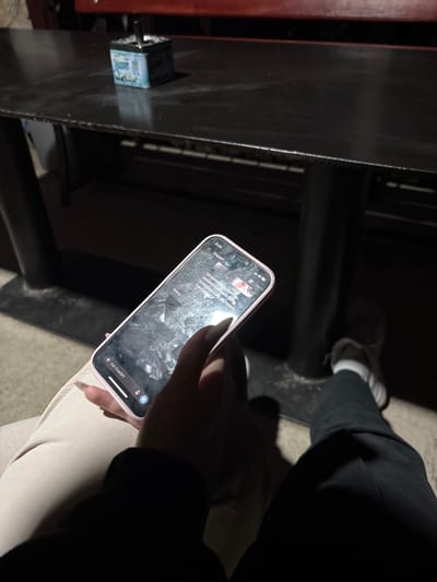 The image shows a close-up of a person's hand holding a pink-cased smartphone with the ChatGPT interface displayed on the screen. The phone is positioned over a person's lap, who is wearing light-colored pants. Another person, wearing dark pants and white sneakers, is visible in the lower right portion of the frame. They are seated at a dark-colored table. On the table, an ashtray with a cigarette is visible. The environment appears to be outdoors, possibly a patio or seating area, with dark, possibly metallic, table legs and a red element suggesting a larger structure or awning above. The lighting is dim, indicating it is likely evening or night. There is no specific indication of the city of Pechinska, Bulgaria, in the image itself.