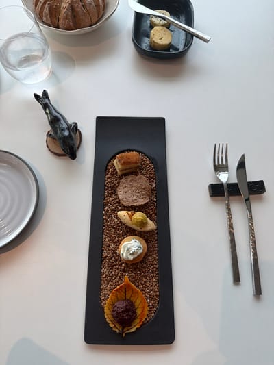 A overhead shot of a white table shows a meal setting in La Clarté, France. The meal includes a circular loaf of bread in a silver bowl, a glass of water, a small black figurine of a seal on a wooden base, a black rectangular plate filled with buckwheat and topped with small bites, a fork and knife resting on a black holder, and a white plate on the left side of the frame. The small bites on the black plate are arranged vertically and consist of a piece of toast with a golden crust, a circular cracker topped with a dark spread, a cracker with a yellow spread, a cracker topped with white cream and herbs, and a decorative leaf with a dark topping. The setting appears to be a restaurant.