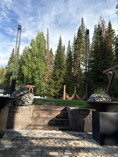 The image depicts an outdoor patio area with a stone staircase leading to a grassy lawn, surrounded by a dense forest of coniferous and deciduous trees. Two modern, tall, black metal lamp posts stand on either side of the staircase. In the foreground, a cobblestone patio is visible, with large, dark, cubic planters and two spherical gabion planters filled with stones and vegetation. A hammock is strung between two wooden posts in the middle ground. In the background, several white spherical objects are scattered on the lawn. The sky is blue with scattered white clouds. The scene appears to be a serene and well-maintained outdoor space, likely part of a residential property or a resort, located in a natural, forested environment.