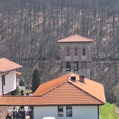The image shows a stone bell tower with a red-tiled roof standing behind a building also with a red-tiled roof and white walls. Both buildings are situated in Gigintsi, Bulgaria. The tower has arched windows and decorative stone banding. In the foreground, a gathering of people is visible near the building. The background is filled with a dense forest of leafless trees.