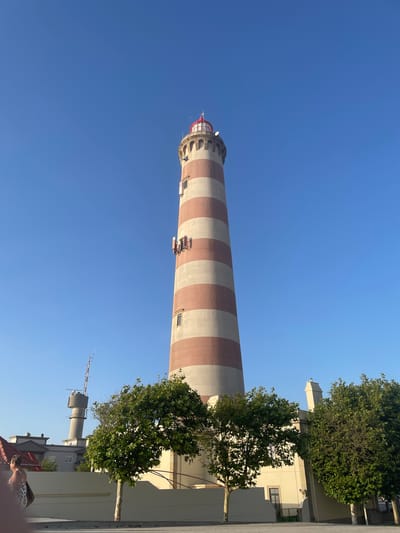 The image depicts the Barra Lighthouse (Farol da Barra) in Gafanha da Nazaré, Portugal, under a clear blue sky. The lighthouse is a tall, cylindrical structure with alternating bands of light beige and reddish-brown. It is topped with a red lantern room and antenna. Mounted on the side of the lighthouse are several antennas or communication devices. In the background, a smaller, cylindrical tower with an antenna and radar equipment is visible, along with the roofs of some buildings. In the foreground, there are several leafy green trees, and a low, light-colored wall or barrier. To the far left, partially visible, are two individuals walking. The overall scene is that of a coastal area with prominent maritime infrastructure.