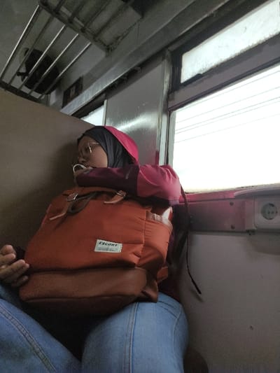 The image shows a person resting inside what appears to be a train or similar public transport vehicle, likely in Kecamatan Beji, Indonesia. The primary subject is a person seen from the chest up, wearing a red hooded top or jacket over a dark head covering and glasses. Their eyes are closed or partially closed, suggesting they are sleeping or resting during travel.
A prominent orange-brown backpack is positioned in front of the person, possibly supporting their head or acting as a lap item. The backpack features a white rectangular label with the word "ESCORT" clearly visible in black text. To the right, part of another person's leg or lap, clad in blue denim jeans, is visible.
The setting is an interior space with bright light coming from a large window or translucent panel in the upper left, indicating it is likely daytime. A power outlet or similar fixture is mounted on the wall in the upper right. In the lower left, a metal grate or rack structure is visible, consistent with the interior design of a public transport carriage. The scene conveys a moment of quiet repose during a journey.