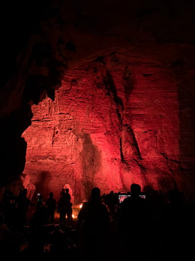 A group of silhouetted individuals stands in the foreground of a dimly lit cave, illuminated by a vibrant red light. The cave walls are rough and textured, with visible striations and formations, likely limestone. The red lighting casts dramatic shadows and highlights on the rock surfaces, creating an ethereal atmosphere. Some people are holding up phones, possibly taking pictures or videos. The environment appears to be a natural cave, possibly a tourist attraction, given the presence of people and lighting. The location is identified as Gollagutta, India.