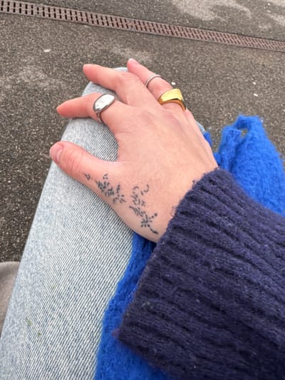 A person's hand with a floral tattoo, adorned with three rings, rests on their denim-clad thigh. The hand is positioned between the person's dark blue sweater sleeve and a vibrant blue, possibly knitted, garment. The rings include a gold square ring, a silver ring with small indentations, and a silver signet ring. The scene is captured outdoors, with a section of pavement visible in the background.