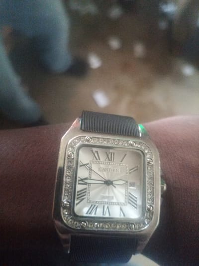 A close-up shot captures a wrist wearing a square-faced Cartier watch. The watch features a silver-toned case adorned with small, glittering stones around its bezel. The watch face is white with Roman numerals, and the brand name "CARTIER" is visible above the word "QUARTZ". The watch band appears to be black and textured. The surrounding environment is blurred, with hints of dark and light tones suggesting an indoor setting, potentially a residential area within Zot Shen, Nigeria. The image focuses on the watch, indicating it is the primary subject of interest.