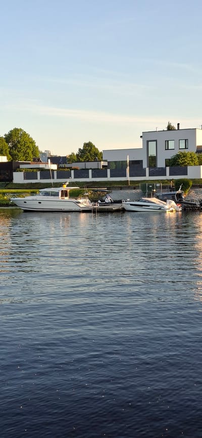 This is a photograph of a body of water, likely a river or bay, with several boats docked along the shore. The water is calm, with ripples reflecting the sunlight. On the shore, there is a modern building with large windows and a dark-colored roof. Several trees are visible behind the building. In the foreground, three boats are docked: a white Nimbus Nova motor yacht, a smaller white speedboat, and a portion of another vessel to the far right. The Nimbus Nova has the text "NIMBUS NOVA" visible on its hull. The scene appears to be located in Pärnu linn, Estonia.