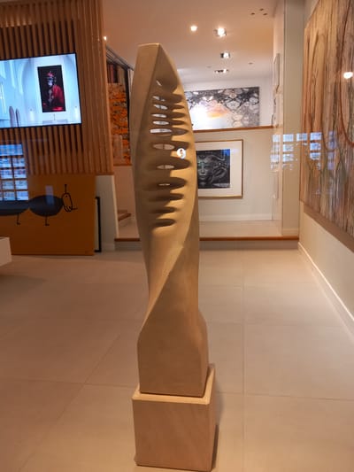 The image depicts a sculpture, likely of sandstone or a similar material, with a twisted, aerodynamic shape featuring repetitive, rounded cutouts along its length. It rests on a two-tiered, rectangular plinth of the same material. The sculpture is positioned indoors, within what appears to be an art gallery or museum setting.
The background includes a series of framed artworks hanging on the walls. Visible among them are a large abstract painting with swirling colors and a more realistic portrait in a gold frame. The lighting is ambient, casting soft shadows.
Below the sculpture and behind it is a wooden slat wall concealing a television screen that displays a portrait of a man in red clothing. Adjacent to the television is a colorful abstract artwork.
This image was taken in the city of Port D'andratx, Spain.