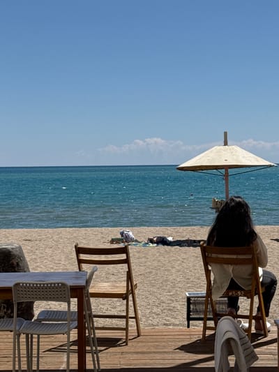 The image depicts a sunny day at a beach in Koshkol', Kyrgyzstan. In the foreground, a wooden deck features a table with white plastic chairs. To the right, a woman with long dark hair sits in a wooden folding chair, facing the water. She is wearing a light-colored shirt and dark pants. Next to her chair, a small black box-like object is visible on the deck. Beyond the deck, a sandy beach stretches towards the turquoise waters of a lake, with a large weathered rock on the left side of the sand. Further up the beach, an umbrella stands planted in the sand, and two individuals are lying down on a blanket. The sky is a clear, bright blue with a few wispy clouds.
