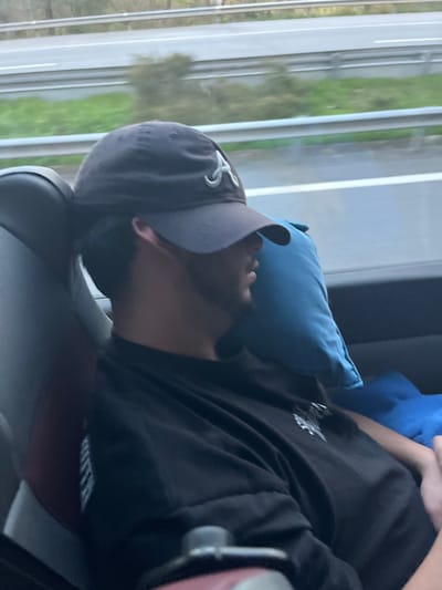 A male individual is depicted seated within a moving vehicle, observed from a slightly elevated, oblique angle. He is wearing a dark baseball cap featuring a white stylized "A" logo on the front and a dark short-sleeved t-shirt. His head is tilted to the right, resting against a light blue neck pillow positioned between his head/neck and the vehicle's window. Dark hair is visible from beneath the cap, and he has a visible dark beard or heavy stubble. The vehicle interior shows a grey upholstered seat. Outside the window, a multi-lane asphalt highway is visible, separated from a strip of green vegetation by a grey metal guardrail. The exterior environment appears blurred, indicating the vehicle's motion. The specific location within the city of Leiria, Portugal, cannot be determined from the visual information provided in the image, as only a generic highway scene is depicted.