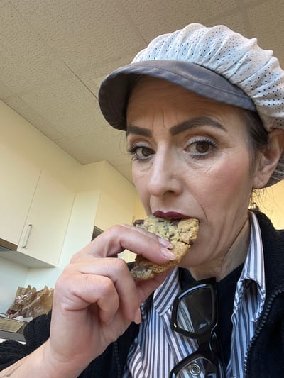 A person, likely female, is shown from a low angle, holding a partially eaten chocolate chip cookie close to her mouth. She has brown eyes, dark eyebrows, and is wearing a white surgical cap with a grey brim. The person appears to be wearing a striped shirt underneath a black jacket. She is wearing black glasses. The background includes a tiled ceiling, white cabinets, and what appears to be a stack of brown paper bags. The location is purported to be Mensdorf, Luxembourg, but this is not visually verifiable from the image.
