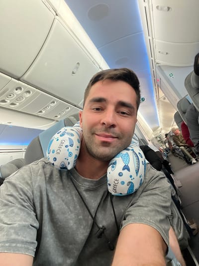 The scene depicts the interior of a commercial aircraft cabin. A male individual, appearing to be in his late 20s to early 30s, is centered in a self-portrait. He has short dark hair, light facial hair, and a slight smile, looking directly into the camera. He wears a grey short-sleeved t-shirt and a light blue and white neck pillow adorned with blue fish patterns and the word "GREECE" repeated multiple times. A black cord necklace with a small black pendant is visible around his neck. The aircraft cabin features white overhead compartments, passenger service units, and overhead lighting, including a blue accent light strip along the ceiling. Several other passengers are partially visible further down the aisle, seated in their respective seats. The context of the "GREECE" themed pillow and the specific prompt request places this travel event as relevant to the Paiania Municipal Unit, Greece, likely pertaining to Athens International Airport.