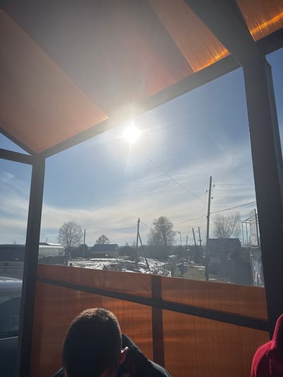 View through a framed window. The foreground shows the corner of a building or structure with orange siding and dark grey framework. The sun shines through the pane, creating a bright glare. The background is the exterior view of the city of Gory, Russia, with a blue sky, light clouds, and electric poles with lines. Buildings and trees line the street. A person's head is partially visible in the lower right corner, and part of their red clothing is in the upper right corner.