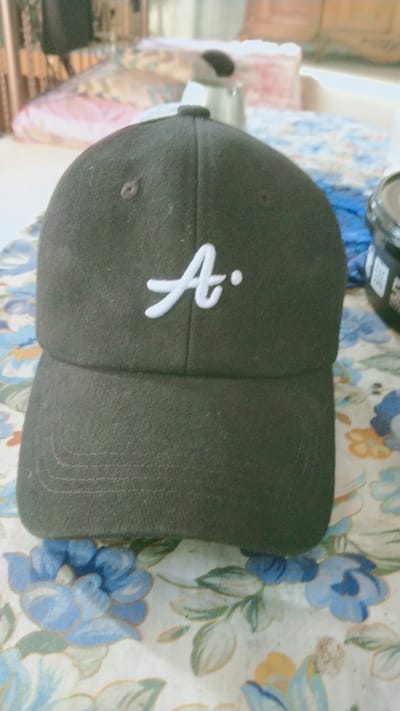 A dark green baseball cap with a white embroidered "A" is centered in the frame, lying on a table covered with a floral-patterned cloth. The cap has visible stitching on the brim and a button on top. In the background, out of focus, are what appear to be furniture and possibly other objects, suggesting an indoor setting. The lighting is soft, and the overall scene is casual. The specific location of Ketoyan, Indonesia, is not visually determinable from the image.
