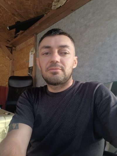 A medium shot selfie is captured from a low angle, showing a Caucasian man with short, dark hair and stubble. He is wearing a dark, short-sleeved t-shirt with a textured pattern. The man's left arm, visible in the foreground, has a tattoo. He appears to be indoors, with a wooden ceiling and walls constructed from panels. Behind him, a black office chair is visible. To the left, a table with a patterned tablecloth is partially shown, with a lighter object resting on it. The background wall is a mottled gray. The scene is lit by natural light, casting shadows. The image was taken in Útušice, Czechia.