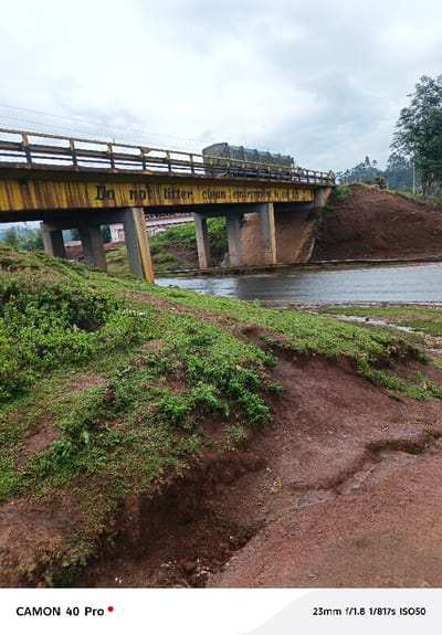 The image captures an outdoor scene featuring a yellow bridge spanning over a wet, dark road. The sky is overcast and light gray, suggesting a cloudy day or recent rain.

The prominent feature is a bridge, painted a distinct yellow color, with a railing along its top edge. Black text is painted onto the side of the bridge, reading: "Do not litter clean environment is our life". The text appears to be hand-painted or stenciled. Several concrete pillars support the bridge. A large truck, possibly covered with a dark tarp, is visible on the bridge, moving from left to right.

Below the bridge, a wet road stretches across the frame, reflecting the dull light from the sky. To the left and in the foreground, there is a significant amount of reddish-brown earth, possibly mud, with patches of vibrant green grass and small plants growing in clumps. The terrain in the foreground is uneven, showing some depressions and possibly water runoff paths in the dirt. On the right side of the road, the ground rises steeply, covered in more reddish-brown earth and some vegetation, leading up to the level of the bridge. In the distant background, beyond the bridge and the rising terrain, some blurry structures and more trees are visible, indicating a more developed or treed area.

The overall impression is one of a functional rural or semi-urban landscape, with clear instructions for environmental consciousness displayed on the bridge. The wet conditions suggest recent precipitation.

Visible text on the image includes:
- On the bridge: "Do not litter clean environment is our life"
- In the bottom left corner: "CAMON 40 Pro"
- In the bottom right corner: "23mm f/1.8 1/817s ISO50"