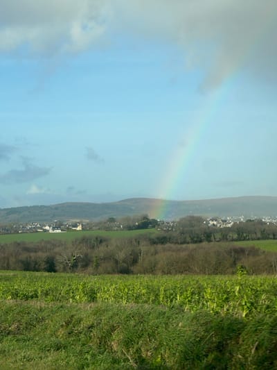 The image displays a rural landscape near Plonévez-Porzay, France, under a light blue sky with scattered grey and white clouds. A distinct rainbow is visible, arcing from the upper right quadrant of the sky down towards the mid-left horizon. The immediate foreground consists of a strip of green grass, transitioning into a wider field of vibrant green crops or tall plants occupying the lower third of the frame. Beyond this field, a band of leafless deciduous trees with visible branches stretches horizontally. Further into the background, a village identified as Plonévez-Porzay is situated, comprising multiple structures with light-colored walls and dark roofs. A prominent, slender spire, likely belonging to a church, is visible within the village on the left side. Rolling hills, covered in vegetation, form the distant background, extending to the horizon. No individuals, animals, or active subjects are present in the scene.